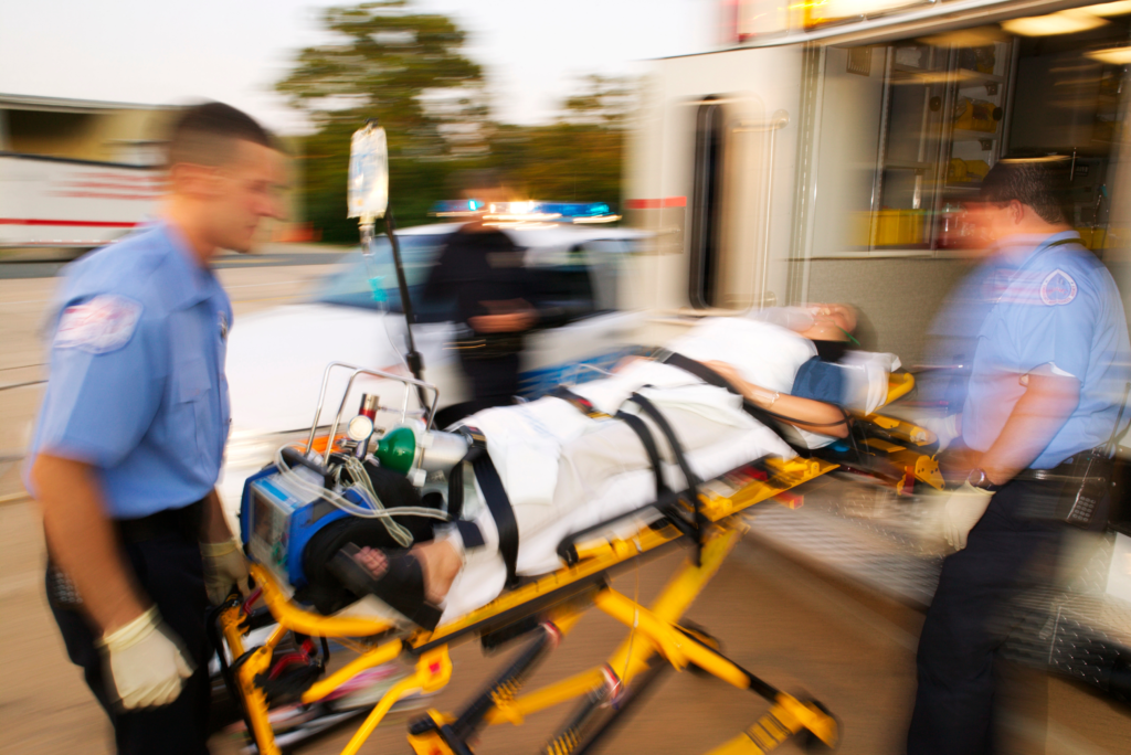 photo in motion of an emergency where two EMS workers are wheeling a patient into the back of the ambulance truck with a cop featured in the background.